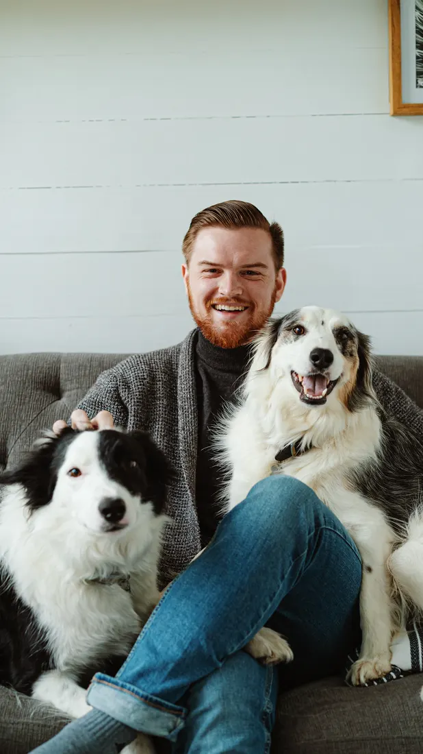 Un hombre sonriente sentado en un sofá con dos perros felices